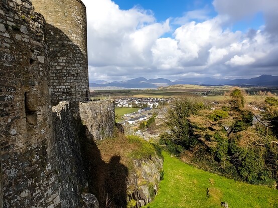Looking past the old stone outer walls of Harlech castle over a flat plain with a small wood of mixed trees and a few streets of modern, mostly white, houses. In the distance a small strip of water can be seen before the mountains of Eryri rise in the distance. The sky is a mixture of thick, fluffy, white and grey clouds with a few patches of blue sky 