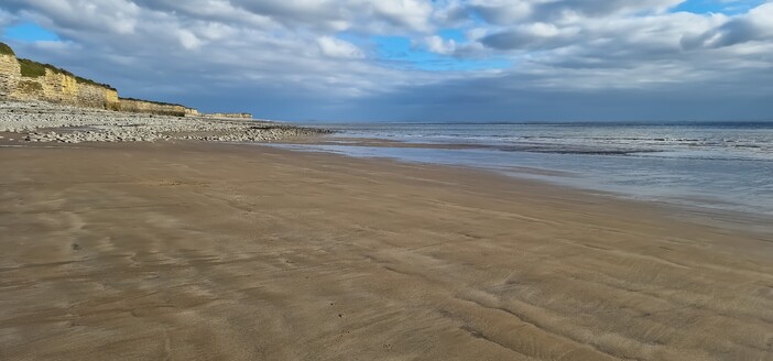 looking down a beach, the foreground is sand but further on it is covered in large pebbles. To the left are high, yellow sandstone cliffs, topped with grass. To the right a calm receding sea breaks gently on the beach, and above a thick layer of white and grey clouds is punctuated by patches of blue sky