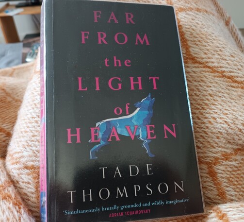 A paperback copy of the novel, Far from the Light of Heaven by Tade Thompson. The cover shows a blue image of a wolf against a black background.