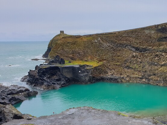 An old quarry right next to the sea, a channel has been made through the narrow wall of rock allowing the sea to flood the quarry forming a deep pool of turquoise water . A small ruined building can be seen on the small strip of land between the sea and the pool and the cliffs rise steeply up behind it and all along the far side of the pool. At the top of the cliff is a small hut like structure looking out tom sea from a headland,  The sky and sea are grey