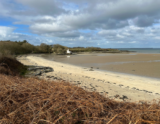 Beach shot at low tide with the sea visible in the distance. Trees and woodland on the left open up onto the beach with a small white derelict building on the rocks in the centre of the photo. Mixed clouds with small patches of blue sky.