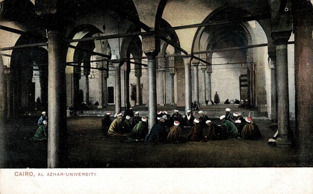 Old postcard showing Al Azhar University, Cairo. The colour card shows a pillared extensive interior space, with groups of men seated in circles on the floor. The men are wearing white and red cloths wrapped around their heads 