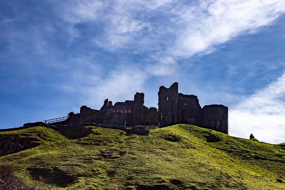 looking up a steep grassy hill to the ruined walls and towers of a castle.  It is seen almost in silhouette against a bright blue sky with some high white wispy clouds