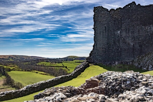 looking past the high outer stone wall of a ruined castle and over lush green farmland  to the horizon.  The sky is blue with high white wispy clouds