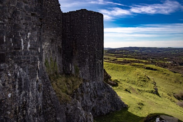 looking past the high outer stone wall and tower of a ruined castle and over lush green farmland  to the horizon.  The sky is blue with high white wispy clouds