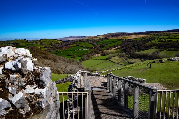 looking from a modern footbridge, giving access to a ruined castle, over rolling hills covered in lush farmland. In the distance the Black Mountains can be seen. Overhead is a cloudless, deep blue sky, marked only by a few vapour trails of distant planes