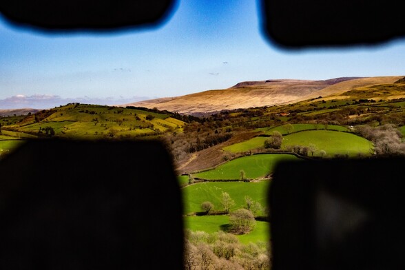 A view through a cross shaped arrow slit in a castle.  Looking out over a patchwork of green farmland and hedgerows to a  distant, barren looking, beige mountain. The sky is pale blue and cloudless apart from a narrow bank of cloud on the horizon