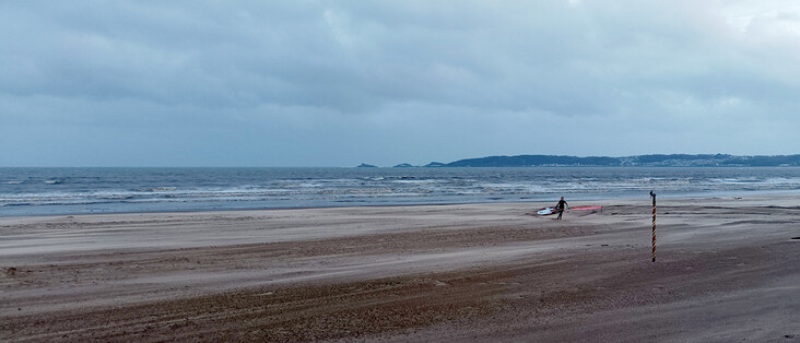 Beach scene: Wide open space. In middle-distance, single windsurfer walks back to shore, waves breaking behind her.