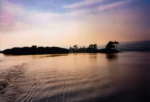 Looking over still water from a boat to a small island seen in silhouette against a twilight sky. The outline of trees can be seen on the island and the wake of the boat on the water