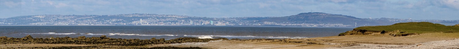 A Panoramic picture looking from the rock, sand and grass of Sker Point over the choppy grey blue water of Swansea Bay to the low hills of Swansea, tightly packed with buildings