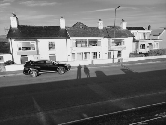 Looking at houses opposite the sea wall in Borth. The shadows of two people standing next to each other on the wall cab be seen on the road and houses.