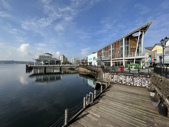 Modern cafes and bars along a decked boardwalk along Cardiff Bay with a calm sea and blue skies. A couple of small piers sticking out into the sea reflected in the water.