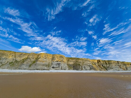 A wide angle shot of a beach. Dark wet sand leads away from the camera to a pebble bank and then to high sheer limestone cliffs. Large rockfalls can be seen along the length of the cliffs. Above the cliffs the sky is a deep blue, marbled with wispy white clouds