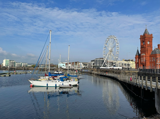 A large red brick building and a ferris wheel along the quay on the right of the picture, with a number of small boats anchored in the water in the centre. Smaller buildings surround the waterfront with a blue sky and mixed white clouds.