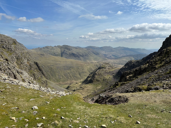 A rocky and grass covered valley heading up between mountains on each side high up towards the summit of Scafell Pike. Lots of smaller mountains are in the distance with a few white clouds and blue skies