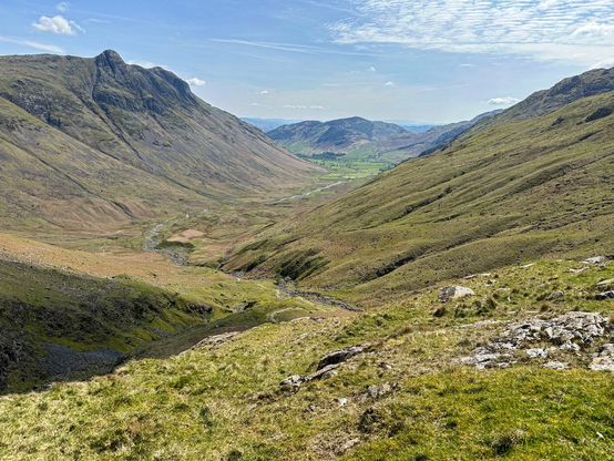 A grass and rocky valley weaves its way between large mountain rains on each side with a rocky path between the hills marking the path towards the summit of Scafell Pike. More hills in the distance covered in rocky outcrops and large patches of grass.