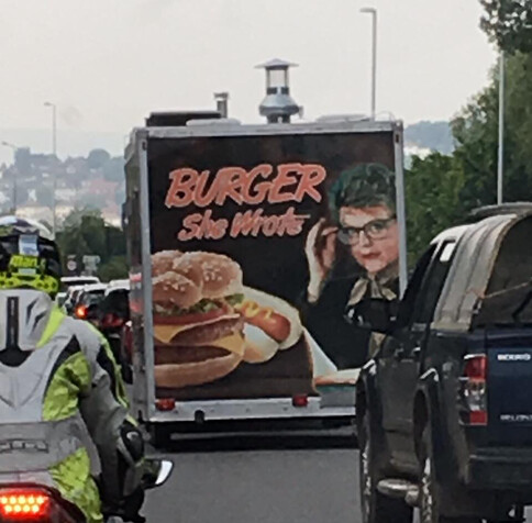 A burger van in a traffic jam, there’s a picture of a burger on the back of the van, next to a picture of Jessica Fletcher and the sign on the van says Burger, She Wrote.