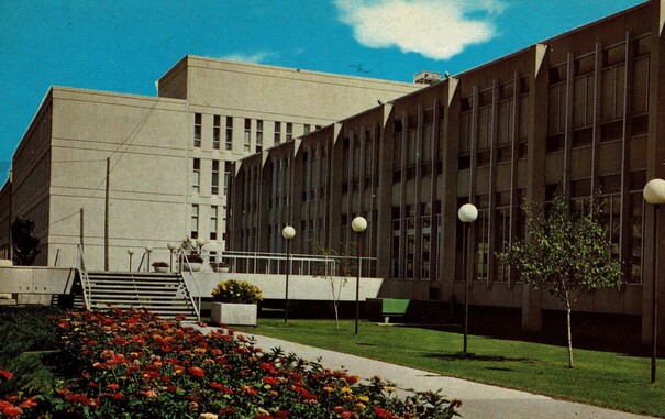 Old postcard showing the University of Manitoba. The colour card shows slab-like concrete buildings, the only features being regular windows, two and four floors high, with blue skies, and a flower bed at the front