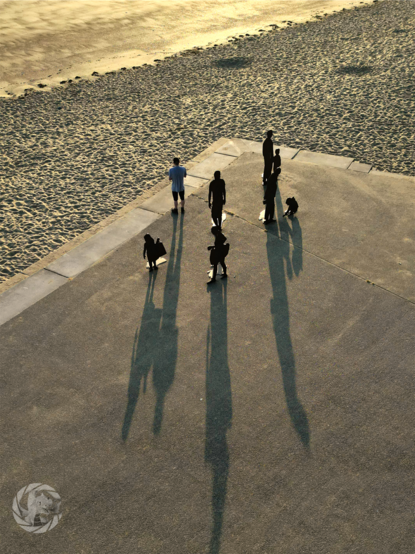 Drone photograph of a number of statues at sunrise, long shadows coming towards the camera. One of the statues is a real person looking out over a sandy beach