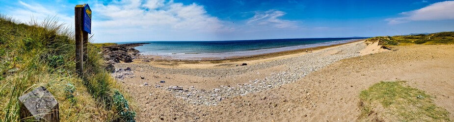 A panoramic view of Sker beach, on the left hand side a blue sign tells us that we are in "Kenfig National Nature Reserve, gwarchodfa natur genedlaethol Cynfig".  Behind the sign, the datk rocks of Sker Point stretch out into a turquoise sea. To the left a sandy beach, backed by pebbles and dunes stretches into the distance