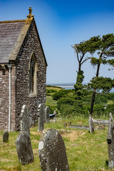 St Madoc's Church in Llanmadoc.

A portrait photo showing the end of a grey stone church with a small cross at the top. Old Gravestones in the foreground with a few tall windblown trees. In the distance a small strip of the blue water of the Lougher Estuary can be seen