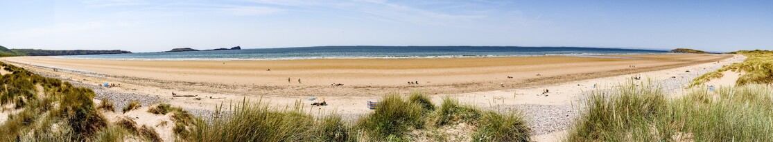 A panoramic picture showing the entire stretch of Rhossilli Bay. 

In the foreground, sand dunes with sparse  Marram grass gives way to a golden sandy beach with just a few sunbathers dotted over it. To the left the land rises to Rhossili village and the "Island" (linked by a tidal causeway), of Worm's Head (so called because it resembles a sleeping Dragon, or Wyrm).  To the right the rocky headland of Burry Holms can be seen