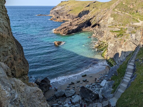A very small, mostly sandy beach inside a rocky cove. There are many steps down to the beach. The sea is clear with dark blue and light blue  colours