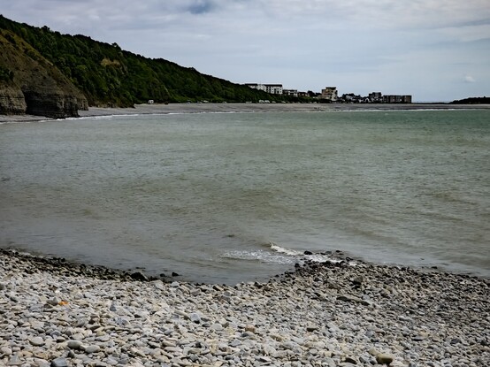 Looking towards "The Knap" from "Porthkerry" Beach.

A pebble beach curves around in a semicircle. Backing the beach are steep cliffs covered in bushes and trees. Directly opposite, over a grey sea, a line of cars are parked at the bottom of the cliffs as they slope down to the houses and flats of The Knap.In the far right of the photo, the edge of the promontory of "Cold Knap Point" can be seen.