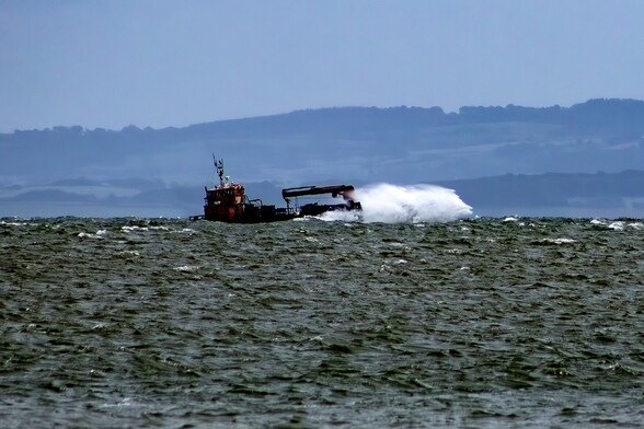 A small ship with a crane arm lying horizontal above it's deck, stryggles through a choppy grey sea. A plume of white spray pushes ahead of it as it breaks through the waves. In the background, the hills of Somerset rise from the Bristol Channel