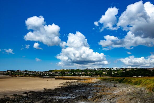Watch House Bay, Barry Island:

looking over dark rocks to a wide beach of golden sand (it was low tide). A harbour breakwater curves out onto the beach. On the opposite shore, the land rises up and is covered in mostly white houses. Above a blue sky is populated with thick white fluffy clouds