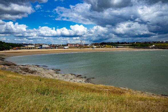 Whitmore Bay, Barry Island:

Looking inland from a grassy promontory, over a flat grey/green sea to a wide sandy beach. Backing the beach is a promenade with a number of shelters and food outlets on it. Further back are the multicoloured "rides" of the amusement park. Above, thick clouds varying from white to dark grey allow the blue sky to peek through in a few areas