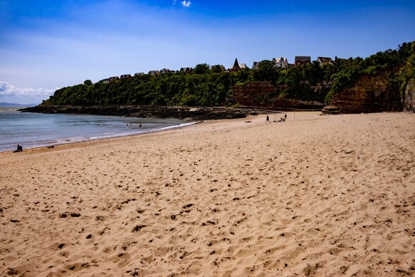 Jackson's Bay, Barry Island:

A wide golden sandy beach, dappled with footprints slopes down to a calm sea. A few people are in the water and sitting on the sand. An abondoned  volleyball net stands part way along the beach. Backing the beach are tall, sheer cliffs, covered in bushes and topped with houses. A clear blue sky sits above the scene