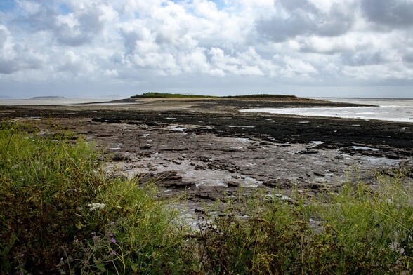Sully Island with Flatholm island in the background:

Looking over a flat rocky beach to a small island, which, as it is low tide, is joined to the beach by a natural causeway. The island slopes gently up from the see on both sides, but dips slightly in the the middle and is covered in green bushes. In the distance, to the left of the first island, another, low, long, flat island can be seen. The sky is grey and filled with thick white and grey clouds, the colour being reflected in a choppy sea