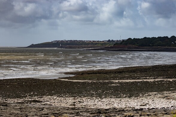 Looking to Nell's Point on Barry Island from Hayes Point on Sully Bay:

A choppy grey sea breaks in small waves a flat rocky beach, much of it covered in black mussels. in the middle distance a rocky promontory, covered in trees sticks out into the bay and behind that the breakwater of Barry docks can be seen with a small white lighthouse at it's end. Further back is Barry Island, topped with houses and at it's far left, the white shape of the Seawatch Centre on Nell's point can just be seen. The sky is grey and filled with thick cloud.
