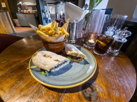 The Treat at the end of the walk:

A pub table, in the foreground is a plate which contains a long soft white bread roll, cut in half and filled with sliced beef steak, and a brown bowl containing thinly cut chips.