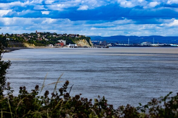 Looking back to Penarth Pier and Cardiff Bay beyond:

An expanse of flat, slate coloured sea stretches out below. A Pier stretches out into it, a few huts set along it's length and a more substantial building sits at it's entrance. Sheer cliffs rise up behind it with the houses of Penarth on top. In the distance the flats and industrial buildings of Cardiff Bay can be seen. A threatening, grey, cloud laden sky sits  above