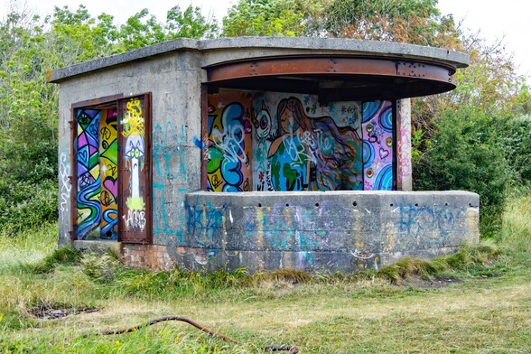 A brightly graffitied WW2 gun emplacement:

A square grey, concrete blockbuilding with a semi-circular bay at the front. It has a rusted metal girder at the top of the semicircular bay. The back of this bay and also metal doors on the side, have been painted with brightly coloured graffitti, some of it depicting ant-war peace messages.