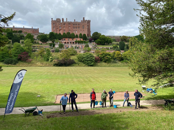 A group of seven people in the foreground, some with digging tools. It is a sunny, but the sky is cloudy. Behind the people is a large lawn, and beyond that - on the crest of a hill - is a large red sandstone castle. There are some ornamental gardens on the slope below the castle. A banner on the left-hand side of the photo reads ‘Clwyd-Powys Archaeological Trust’. 