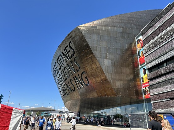 Imposing bronze coloured facade of the Wales Millennium Centre with festival stands in the foreground and bright blue sky 