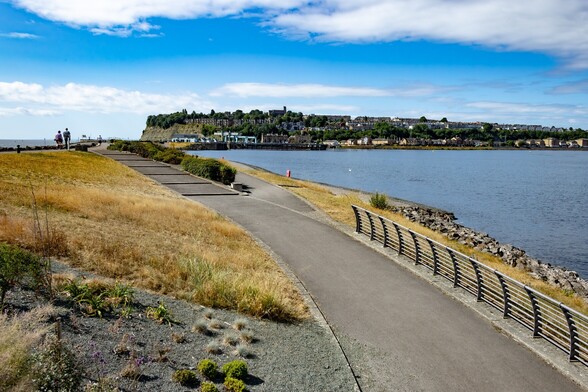 Looking towards Penarth from The Cardiff Barrage:

Two Parallel footpaths, close together but at slightly different levels are joined by another, slightly sloping path. They curve around, following the shape of the barrage and resulting bay. The mechanisms of the sea locks can be seen in the distance and behind them, Penarth. The older houses built on top of a hill above a sheer sea cliff, with newer houses placed around the waterfront. The water is calm and blue, reflecting the pale blue sky above, which is sparsely populated with thin, high white clouds