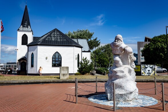 The Norwegian Church and Scott Antartic Memorial:

A small Norwegian style church, it's walls made from white wooden boards and it's roof and spire a faded black sits behind a sculpture depicting Scott man-hauling towards the south Pole. It's surface is made of an irregular mosaic of small white tiles.