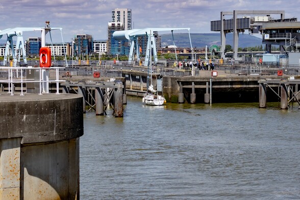 A small yacht moving into one of the sea locks of the Cardiff Barrage:

A white yacht motors into a gap between two concrete walls. It is dwarfed by the size of the lock, only it's mast pokes above the walls. Above, large grey steel arms sit that lever up the roadway to allow boats to pass through. In the background, houses and apartment blocks, some under construction, can be seen on the opposite side of the bay