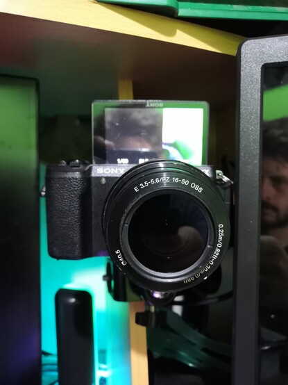 Sony A5100 camera clamped to a vertical wooden strut on the top section of a computer desk, just above level with my computer monitors. My reflection can be seen in the monitors, the camera screen, and the lens filter.