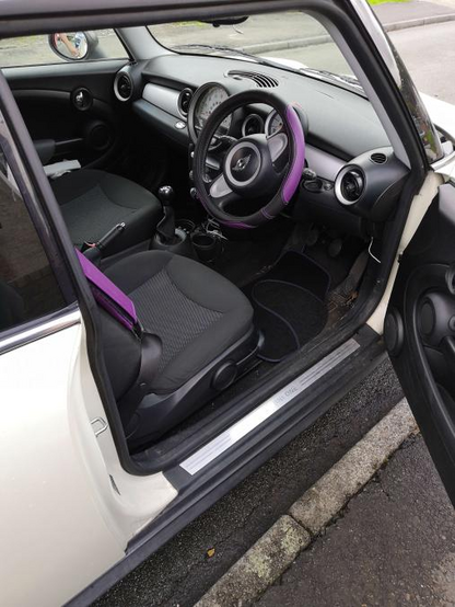 Interior of a Mini One, with a purple padded seatbelt cover, a purple-accented steering wheel cover, and purple-accented floor mats.