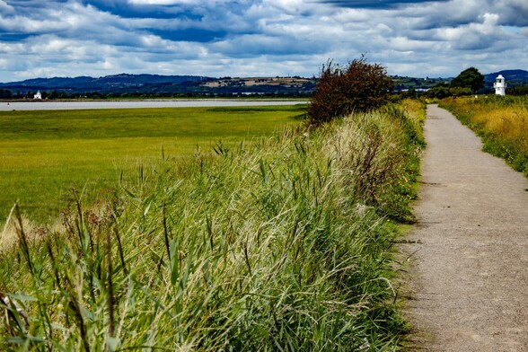 A view along the coast path as it runs parallel to the coastline, through the Newport Wetlands Nature Reserve:

A grey gravel path runs straight ahead towards the white East Usk Lighthouse. Bordered on both sides by low banks of grasses. To the left an expanse of green marshland runs to the edge of the Severn Estuary.. In the distance, and across the mouth of the Usk River, The sqautter West Usk Lighthouse can be seen. Thick grey cloud fills the sky