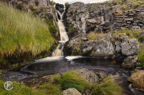 A small stream drains from a peat bog down a 10foot high waterfall. 

A long exposure turns the water silky smooth, and white bubbles have formed swirls in the pool at the bottom of the falls.

Rocks and a dry stone wall on the right hand side, green grasse on the left and immediate foreground