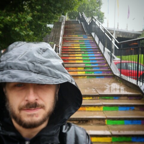 Me wearing a hooded coat in the rain, standing in front of a flight of outdoor steps painted in rainbow colours, leading to the Wyvern Theatre in Swindon.
