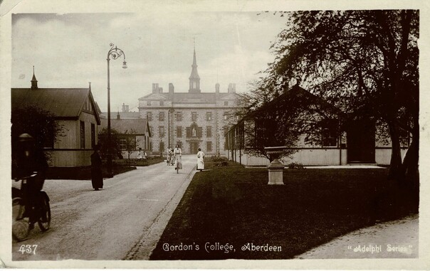 Old postcard showing Gordon’s College, Aberdeen. The monochrome card, in landscape orientation, shows a large building at the end of a drive, with smaller buildings arranged on either side of the drive. There are trees, a couple of pedestrians, and some men and women on bicycles. At the foot of the card is printed in white ink “437 Gordon’s College, Aberdeen, Adelphi Series”