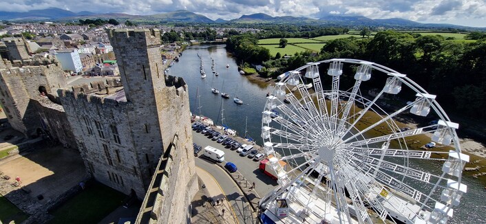 An expansive view from The Eagle Tower at Caernarfon castle. It's a long way up! The castle and it's many towers are to the left. The Arfon river in the middle with little boats on it. Bottom right is a large while Ferris wheel. Green fields to the right. The mountains of Eryri in the distance.
