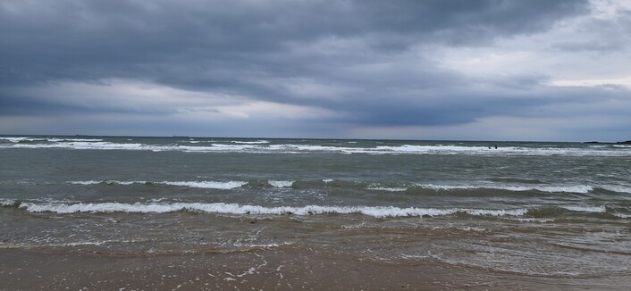 Land, sea and sky. The sky is filled with dark grey cloud with patches of brightness. The sea is looking choppy as the wind starts to pick up. The tops of the waves are highlighted white as they roll over.